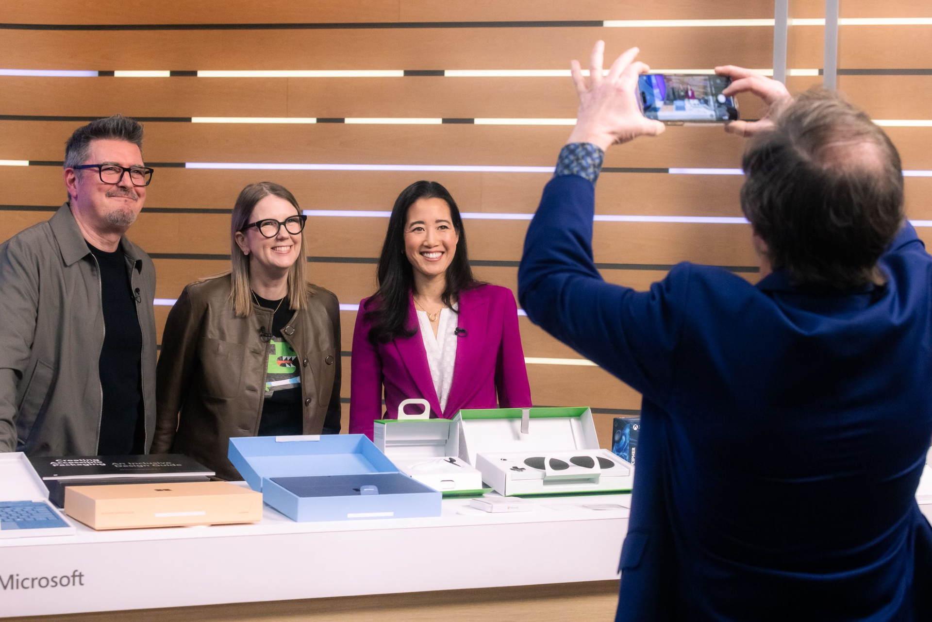 Four people are in the image. Three of them are standing behind a table, and one person is taking a photo of them. The person taking the photo is wearing a blue suit and holding a smartphone. The three people behind the table are smiling. The man on the left is wearing glasses and a brown jacket over a black shirt. The woman in the middle is wearing glasses and a brown jacket over a black shirt with a green design. The woman on the right is wearing a magenta blazer over a white top. The table has several boxes on it, and the word "Microsoft" is visible on the side. The background consists of horizontal wooden panels.
