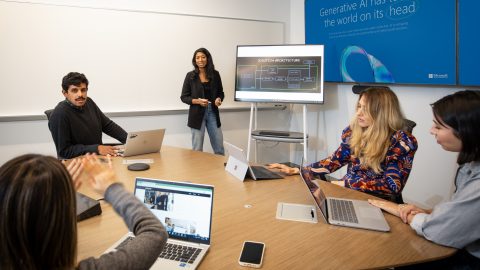 People sitting at a conference table with laptops, with one woman standing in front of a larger screen.