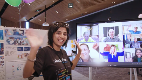 A smiling woman in front of a science fair display and next to a screen showing Microsoft team members.