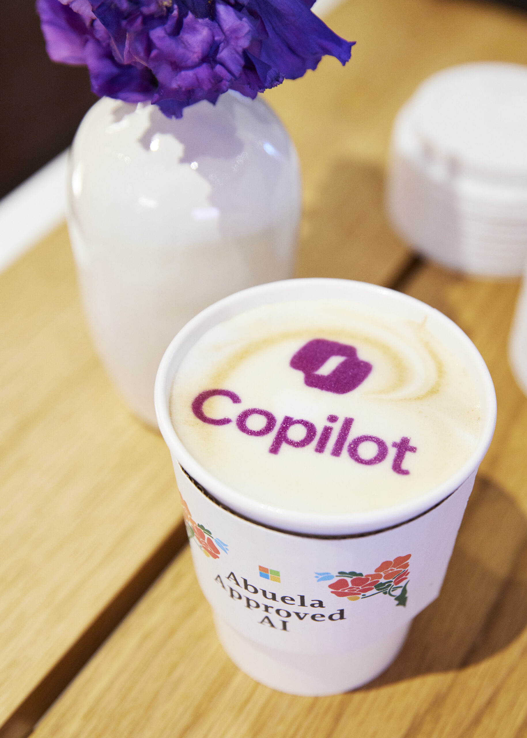 A to-go cup of latte with foam art displaying the Copilot logo, and a cup holder labeled “Abuela Approved AI.
