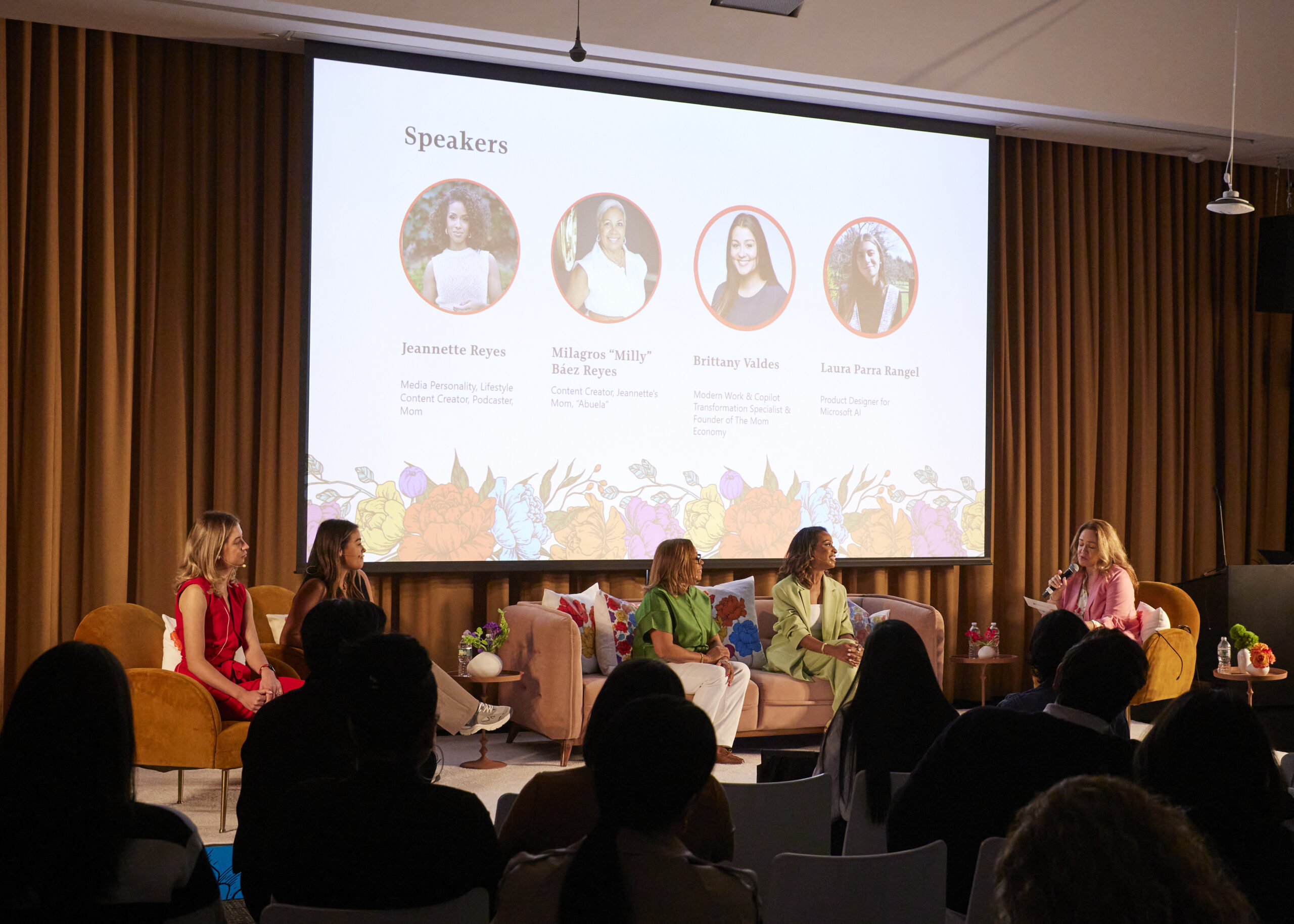 A panel of five women sitting on stage in front of a screen displaying speaker bios at an event.