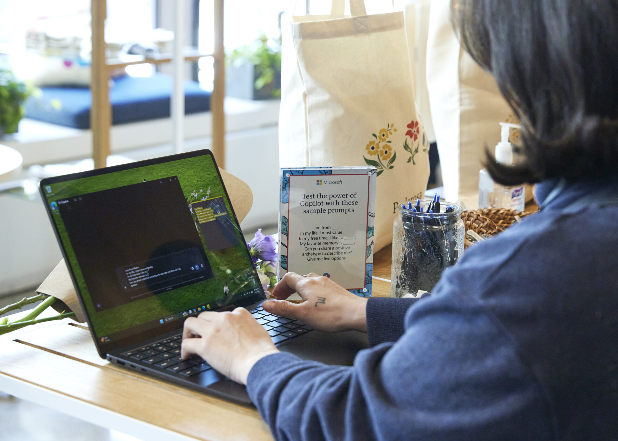 A person typing on a laptop at the Copilot, ¿Quién Soy? station with a small sign, bags, pens, and snacks.