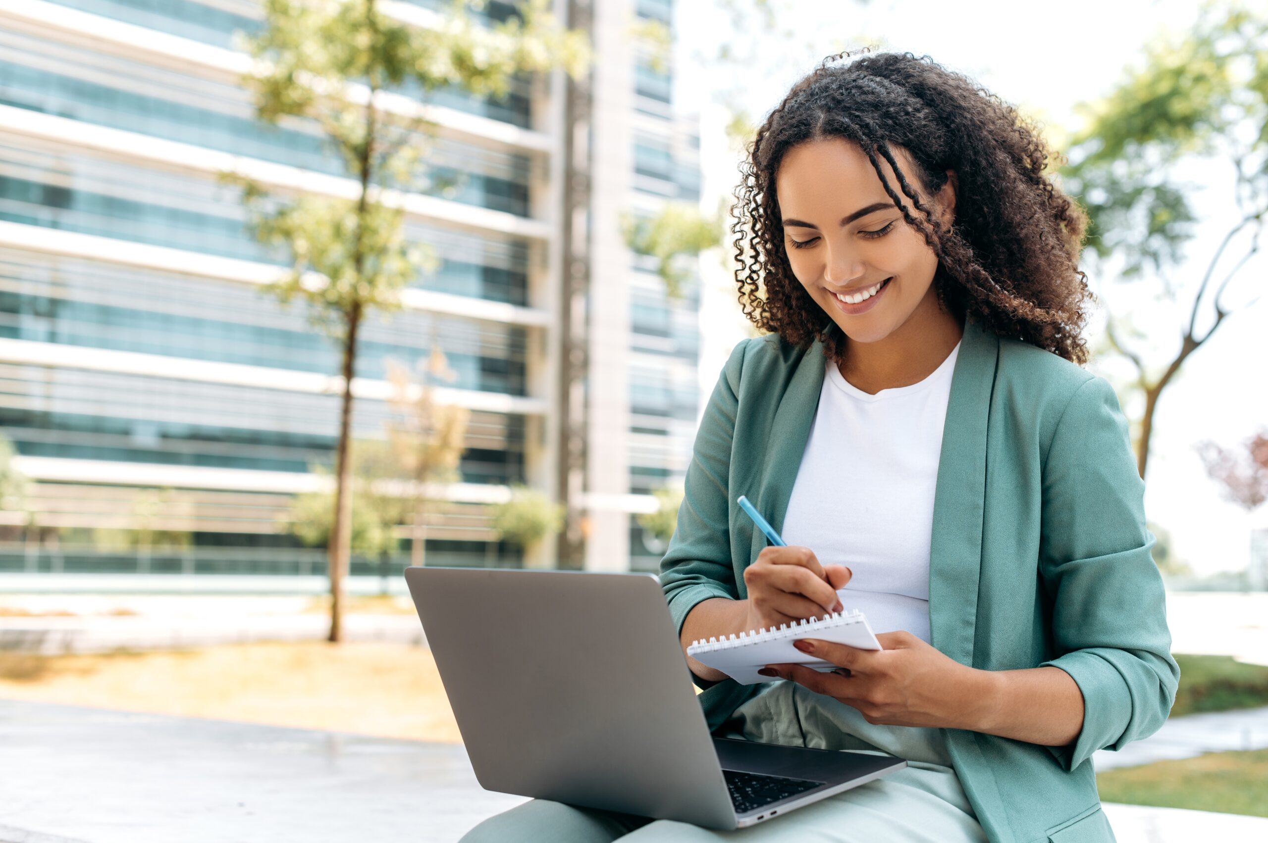 A young woman smiling while writing in a notebook and working on a laptop outdoors.