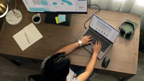 A woman sits at a desk working on a laptop and external monitor