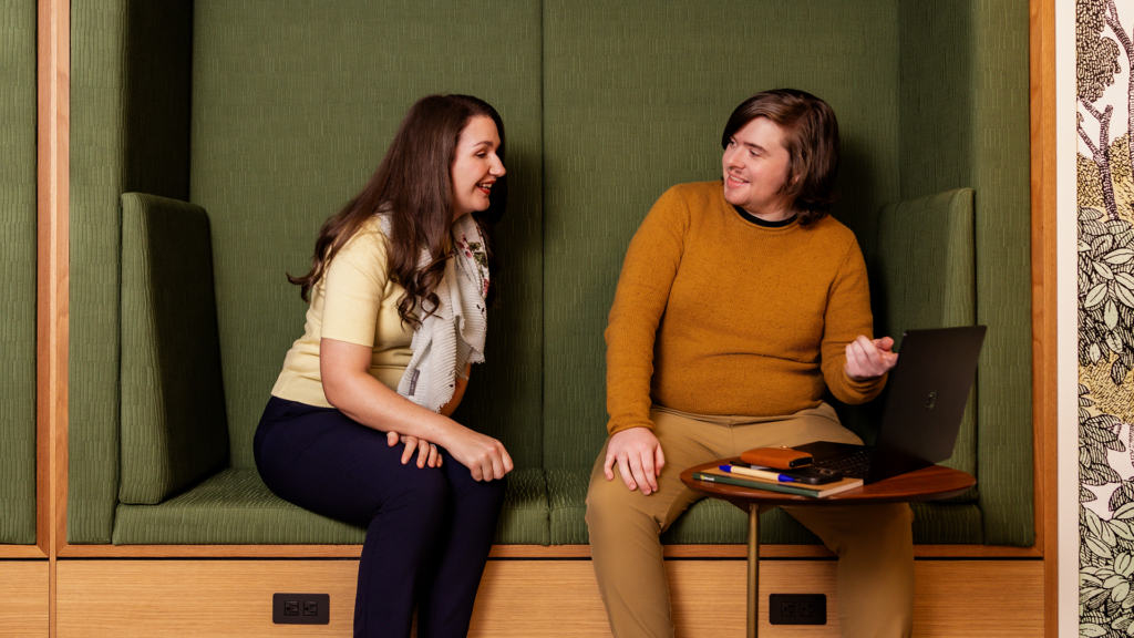 A man and a woman sitting on a bench talking and looking at his laptop in a space filled with natural tones of green, brown and red with warm wood accents. This photo was taken for the Neurodiversity Campaign in a conference room on the Microsoft Redmond Campus.