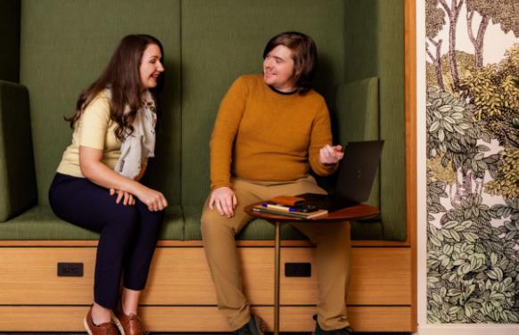An image of two people on a bench referencing a laptop on a small desk nearby.