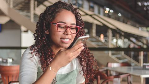 A woman speaks into a cell phone that she holds in front of her face so that she can view the phone’s screen.