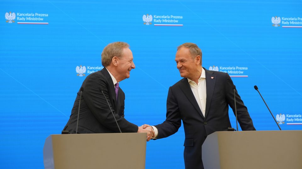 Brad Smith, Vice Chair and President, Microsoft and Donald Tusk, Prime Minister of Poland, in formal attire are shaking hands behind podiums on a stage with a blue background. The background displays the logo and text 'Kancelaria Prezesa Rady Ministrów.