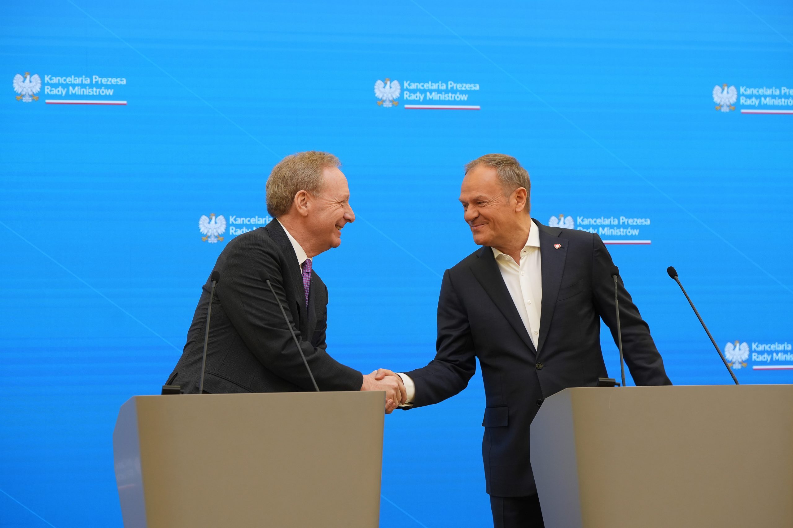 Brad Smith, Vice Chair and President, Microsoft and Donald Tusk, Prime Minister of Poland, in formal attire are shaking hands behind podiums on a stage with a blue background. The background displays the logo and text 'Kancelaria Prezesa Rady Ministrów.