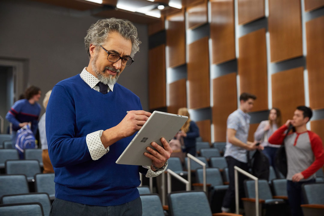 A male university instructor in a lecture hall, using Surface Go to prepare course materials while students file in.