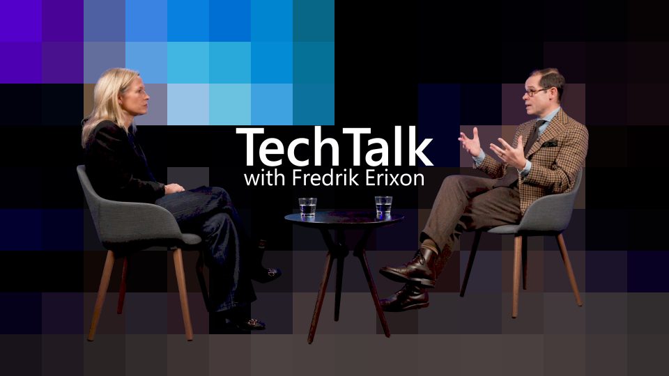 Two individuals are seated on gray chairs facing each other in a studio setting with a black background and pixelated blue and purple squares. The person on the left has long blonde hair and is wearing dark clothing, while the person on the right is wearing a brown checkered blazer, white shirt, and brown pants. A small round table with two glasses of water is placed between them. The text 'TechTalk with Fredrik Erixon' is prominently displayed in white at the center of the image.