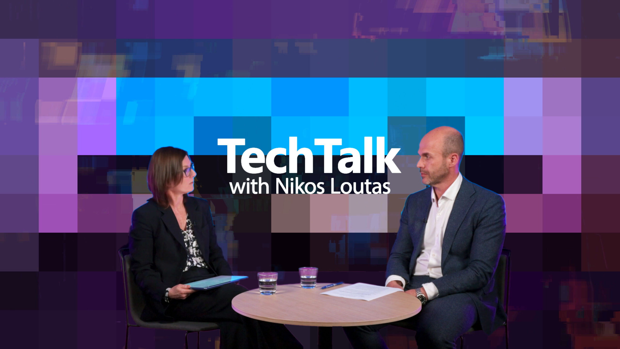 Amy Hogan Burney and Nikos Loutas are seated at a round table with two glasses of water and some papers on it. The background features a colorful pixelated design with shades of blue, purple, and pink.